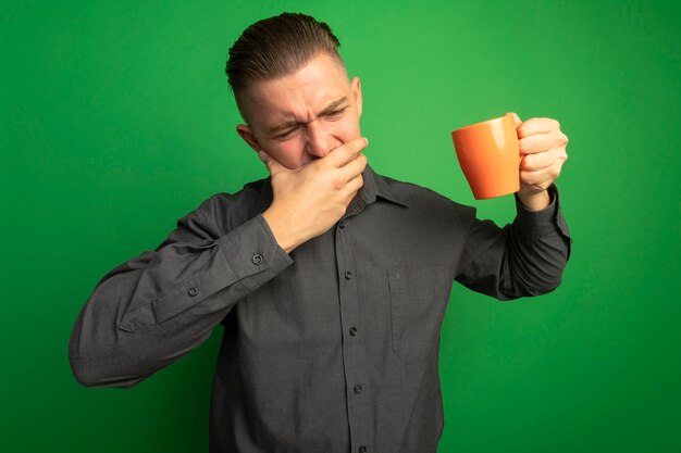 Young handsome man in grey shirt holding orange mug covering mouth with hand feeling discomfort standing over green wall
