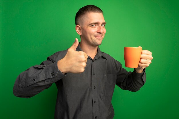 Young handsome man in grey shirt holding orange mug looking at front smiling cheerfully showing thumbs up standing over green wall