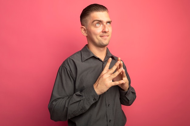 Young handsome man in grey shirt holding palms together smiling slyly looking up standing over pink wall