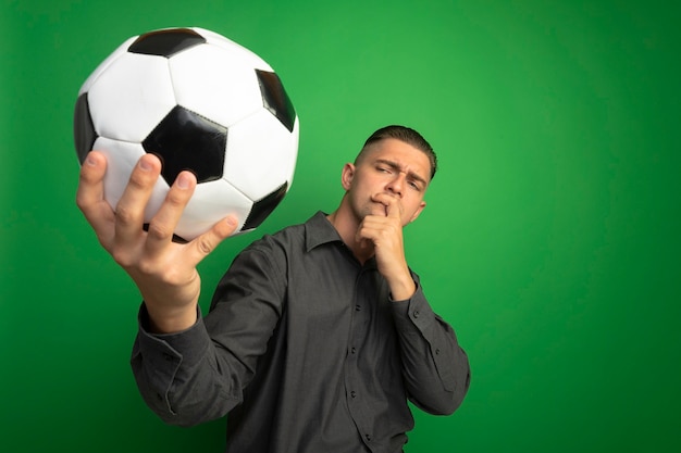 Foto gratuita giovane uomo bello in camicia grigia che tiene pallone da calcio guardandolo incuriosito