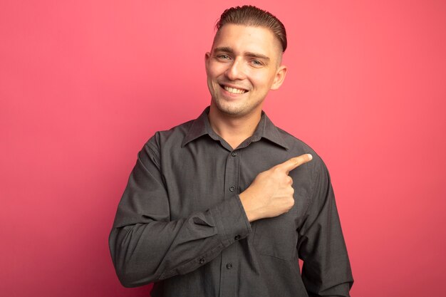 Young handsome man in grey shirt looking at front smiling cheerfully pointing with index finger to the side standing over pink wall