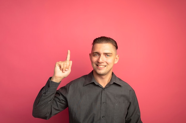 Young handsome man in grey shirt looking at front smiling cheerfully showing index finger standing over pink wall