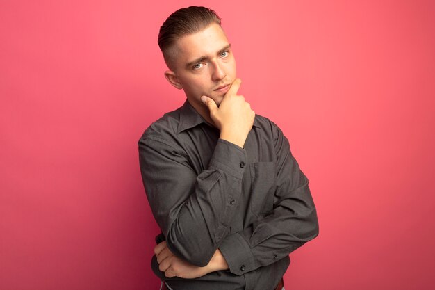 Young handsome man in grey shirt looking at front with hand on his chin thinking standing over pink wall