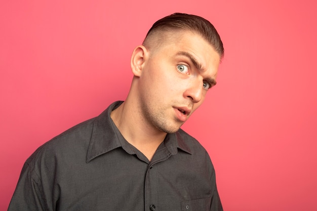 Young handsome man in grey shirt looking at front with skeptic expression standing over pink wall