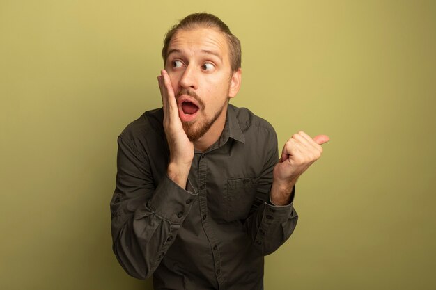 Young handsome man in grey shirt looking surprised pointing with thumb to the side 