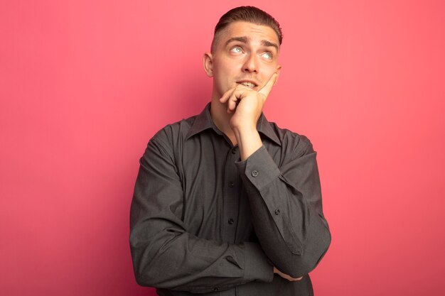 Young handsome man in grey shirt looking up puzzled standing over pink wall