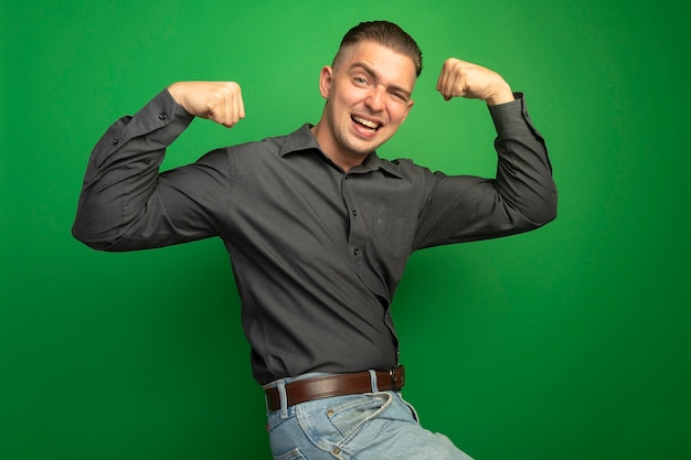 Free photo young handsome man in grey shirt raising fists happy and positive showing biceps and strength winner concept standing over green wall