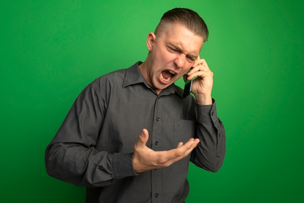 Foto gratuita giovane uomo bello in camicia grigia che grida con espressione aggressiva mentre parla al telefono cellulare in piedi sopra la parete verde