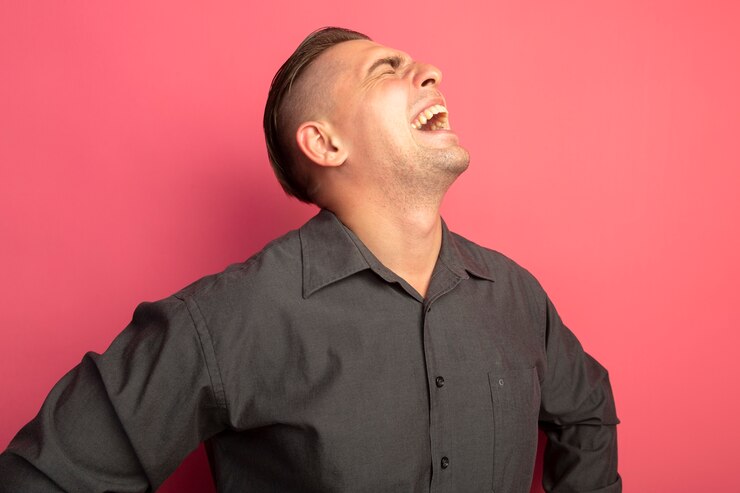 ピンクの壁の上に立っているイライラした表情で叫んでいる灰色のシャツの若いハンサムな男