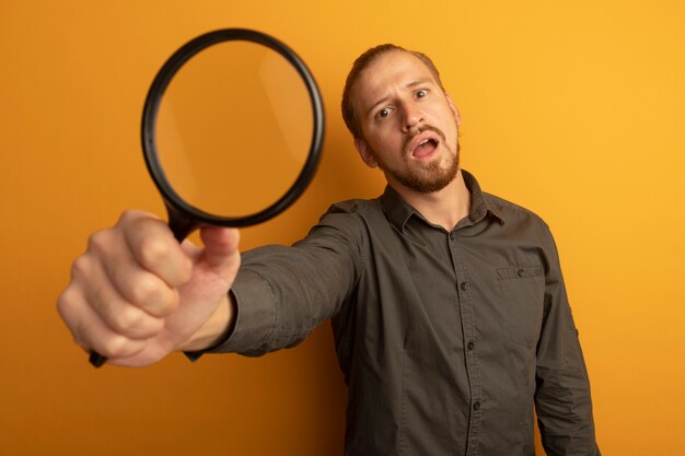 Young handsome man in grey shirt showing magnifying glass confused 