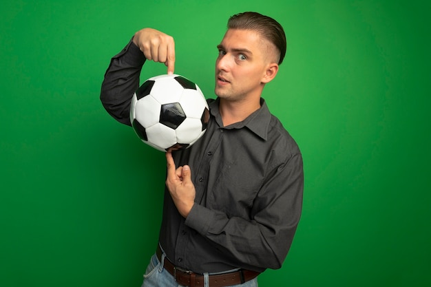 Foto gratuita giovane uomo bello in camicia grigia che mostra il pallone da calcio che punta con il dito indice sorridendo fiducioso in piedi sopra la parete verde