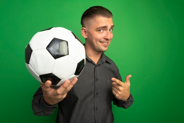 Молодой красавец в серой рубашке показывает футбольный мяч, указывая на него указательным пальцем, улыбаясь со счастливым лицом, стоящим над зеленой стеной
