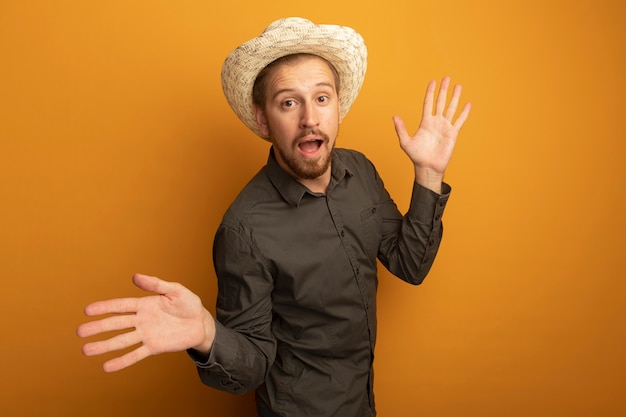 Young handsome man in grey shirt and summer hat confused spreading arms to the sides 