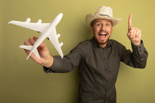 Giovane uomo bello in camicia grigia e cappello estivo che tiene aeroplano giocattolo sorridente felice e positivo che mostra allegramente i pollici in su