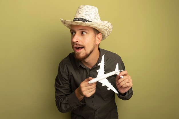 Young handsome man in grey shirt and summer hat holding toy airplane looking aside smiling slyly 