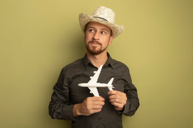 Young handsome man in grey shirt and summer hat holding toy airplane looking aside with shy smile on face 