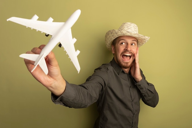 Young handsome man in grey shirt and summer hat holding toy airplane looking at it happy and excited 