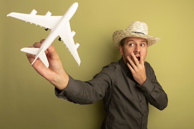 Young handsome man in grey shirt and summer hat holding toy airplane looking at it surprised covering mouth with hand 
