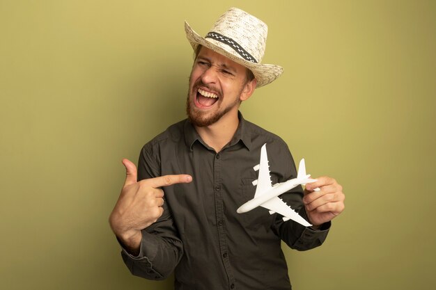 Young handsome man in grey shirt and summer hat holding toy airplane pointing with index finger at it happy and excited 