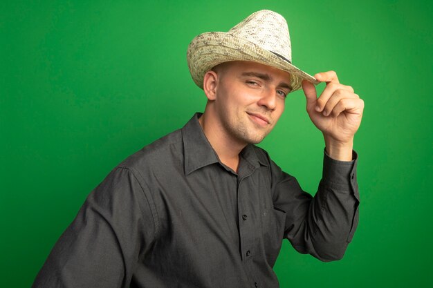 Молодой красивый мужчина в серой рубашке и летней шляпе выглядит уверенно, касаясь его шляпы, улыбаясь, стоя у зеленой стены