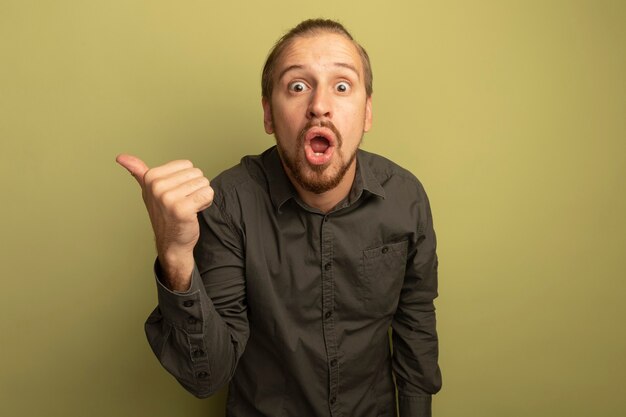 Young handsome man in grey shirt surprised pointing with thumb to the side 