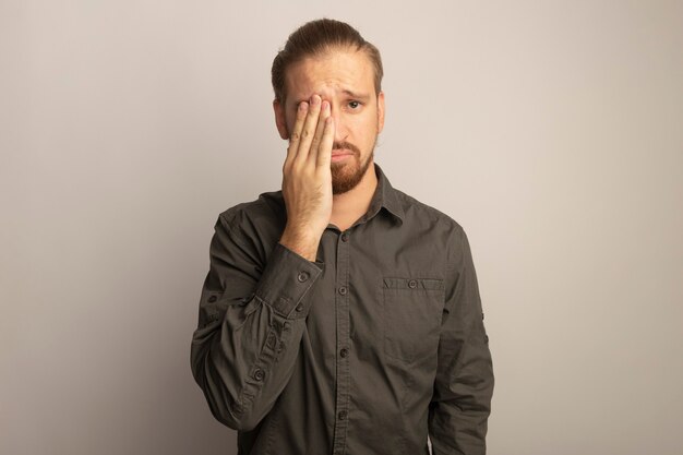 Young handsome man in grey shirt with sad expression covering eye with hand 