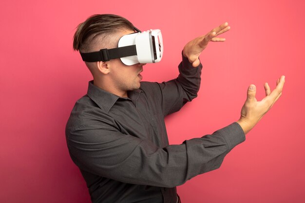 Young handsome man in grey shirt with virtual reality glasses looking aside gesturing with hands standing over pink wall