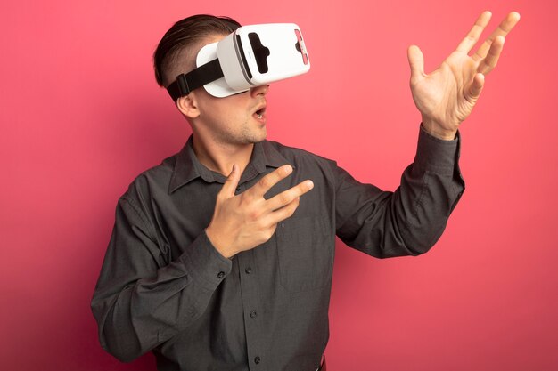 Young handsome man in grey shirt with virtual reality glasses looking aside gesturing with hands standing over pink wall