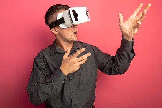 Молодой красавец в серой рубашке с очками виртуальной реальности смотрит в сторону, жестикулируя руками, стоящими над розовой стеной
