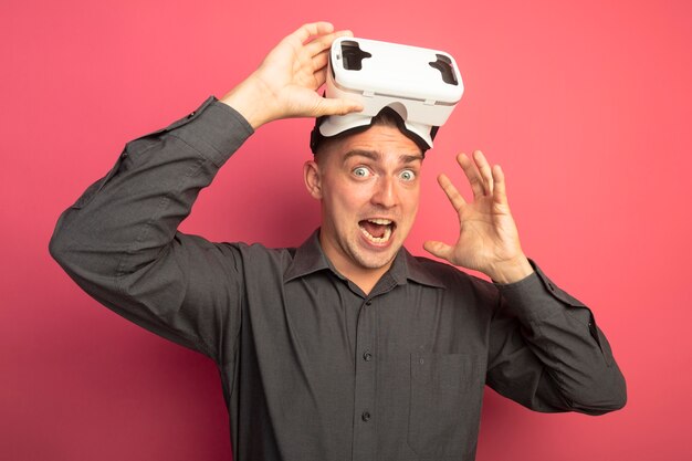 Young handsome man in grey shirt with virtual reality glasses looking at front excited and happy with raised hand standing over pink wall