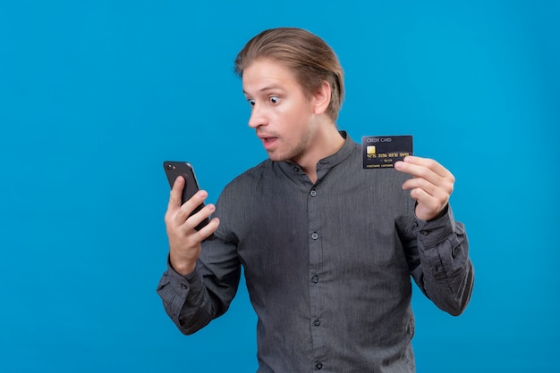 Молодой красивый мужчина держит кредитную карту и смотрит на экран своего смартфона, стоя над синей стеной