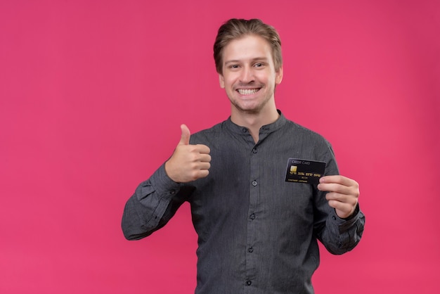 Young handsome man holding credit card showing thumbs up, smiling standing over pink wall