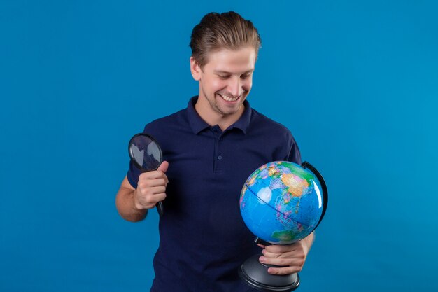 Молодой красивый мужчина держит глобус и увеличительное стекло, глядя на глобус, улыбаясь со счастливым лицом, стоящим на синем фоне