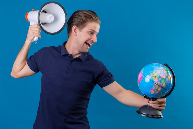 Young handsome man holding globe and megaphone looking playful and happy standing over blue background