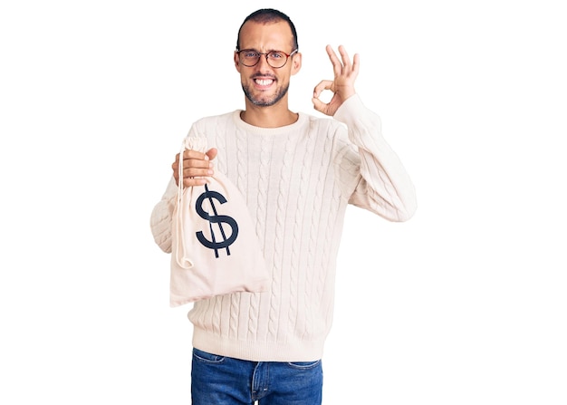 Foto gratuita giovane uomo bello che tiene la borsa dei soldi con il simbolo del dollaro che fa il segno giusto con le dita sorridenti che gesturing amichevole simbolo eccellente