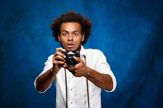 Young handsome man holding old camera over blue surface