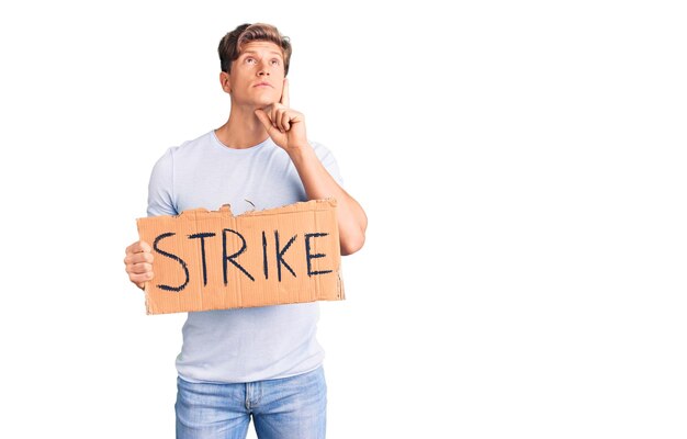 Young handsome man holding strike banner cardboard serious face thinking about question with hand on chin, thoughtful about confusing idea
