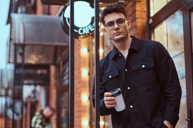 A young handsome man holding a takeaway coffee while standing near a cafe outdoors.