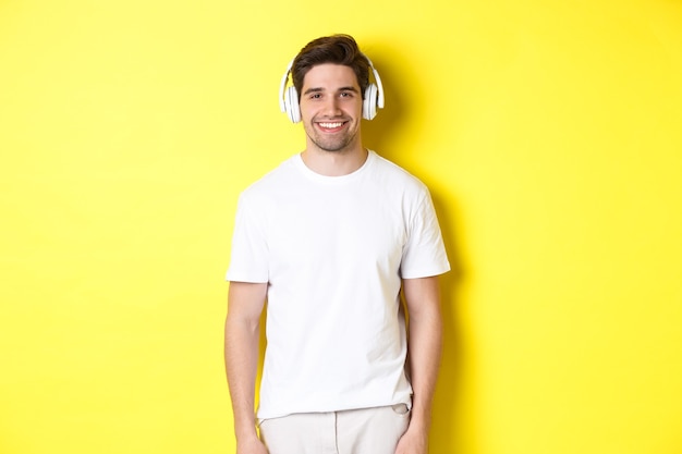 Young handsome man listening music in headphones, wearing earphones and smiling, standing over yellow background.