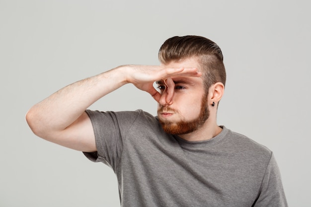 Young handsome man pinching nose over grey wall.