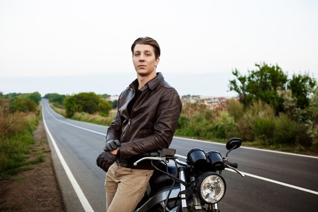 Молодой красавец позирует возле своего мотоцикла на сельской дороге.