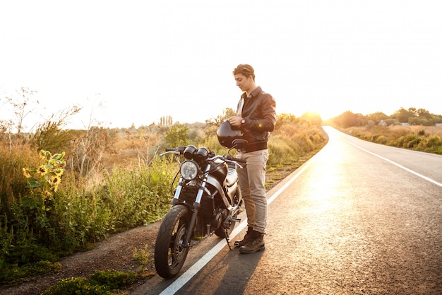 Young handsome man posing near his motorbike at countryside road.