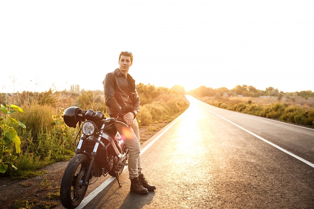 Young handsome man posing near his motorbike at countryside road.