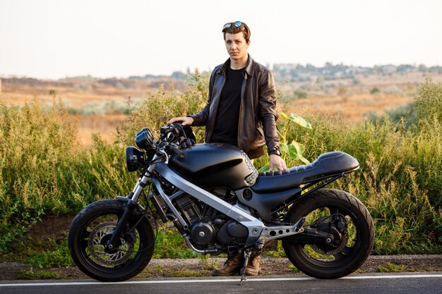 Young handsome man posing near his motorbike at countryside road.