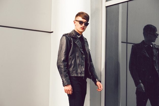 Молодой красавец позирует возле современного бизнес-центра, одетый в стильную кожаную куртку с шипами, черные джинсы и солнцезащитные очки, брутальный вид.