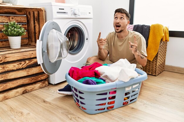 Young handsome man putting dirty laundry into washing machine amazed and surprised looking up and pointing with fingers and raised arms.