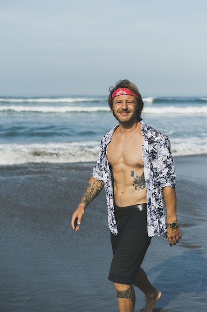 A young handsome man in a red bandana walks along the ocean at the beach.