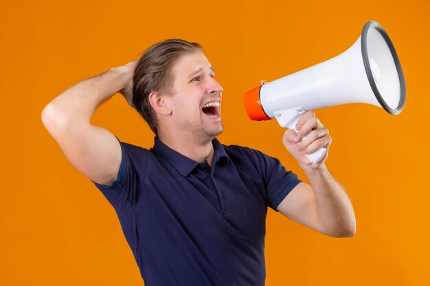 Young handsome man shouting through megaphone exited and surprised standing over orange background