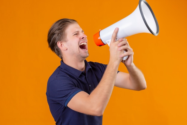 Young handsome man shouting through megaphone exited and surprised standing over orange background