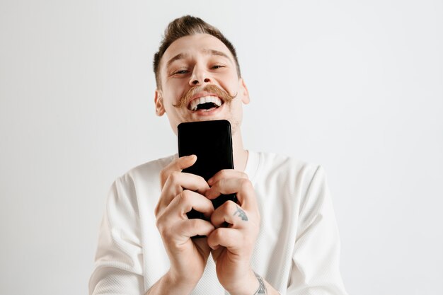 Young handsome man showing smartphone screen over gray background with a surprise face. Human emotions, facial expression concept